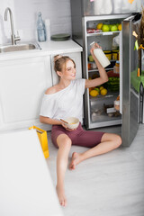 cheerful woman in white t-shirt and shorts holding bottle of milk and bowl with cornflakes while sitting on floor near opened fridge