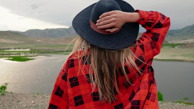 A Woman In A Red Plaid Shirt And Black Hat Against The Backdrop Of The Lake And Mountains. The Wind Plays With The Woman's Long Blond Hair