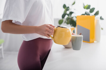 cropped view of young woman pouring tea from teapot in kitchen