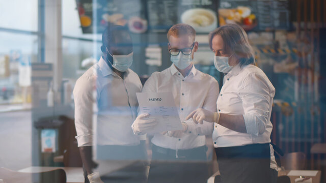 View through window of multiethnic waiters in safety mask and gloves wearing uniform discussing menu