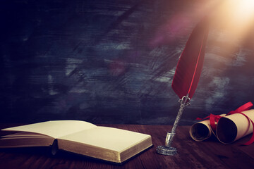 Old feather quill ink pen with inkwell and old books over wooden desk in front of black wall...