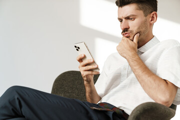 Photo of brooding man using mobile phone while sitting in armchair