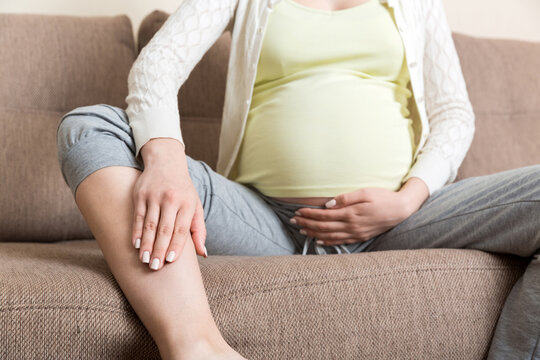 Leg Cramps During Pregnancy. Closeup Of Hands Massaging Swollen Foot While Sitting On Sofa