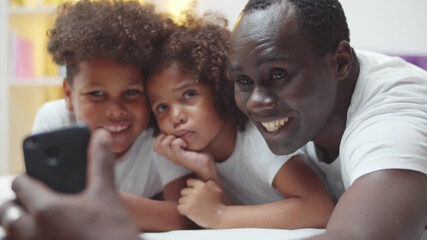 Obraz premium Happy african american father with little son and daughter taking selfie on smartphone at home