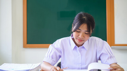 Happy young pretty asian student girl taking homework or reading book alone in classroom with smiling at front green whiteboard, Education learning by myself in white uniform on desk stack of books