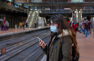 Fototapeta na wymiar en Mujer revisando el teléfono móvil con mascarilla en una estación de tren en Madrid