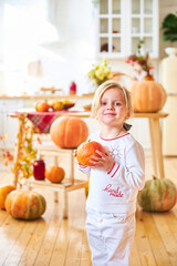 Fototapeta premium A little blonde girl sits on a wooden floor in a white kitchen next to a large orange autumn pumpkin