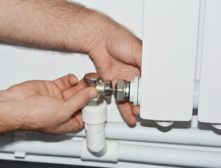 A plumber is connecting chrome fittings, valves of a radiator heater and a plastic central heating pipe installed along the wall.
