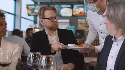 Young bearded man dining with friends returning poor cooked dish to waiter in mask