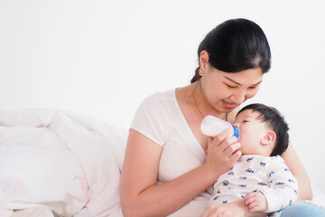 Fototapeta na wymiar Asian mother gently feeding her cute newborn healthy baby with milk bottle in the bedroom at home with pure love and care Mom and child and family lifestyle concept