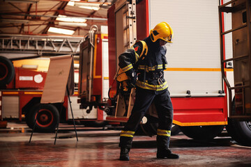 Full length of brave fireman standing in fire station in full protective uniform and preparing for...