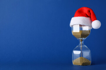 Hourglass and Santa hat on blue background, space for text. Christmas countdown - obrazy, fototapety, plakaty