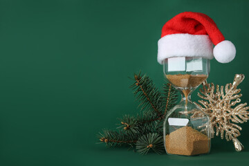 Hourglass with Santa hat and fir tree branches on green background, space for text. Christmas...