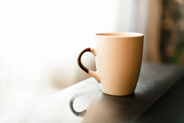 Silhouette of beige ceramic coffee mug placed on dark table near window. Blurred background with reflection of a mug and free space for copy text. Selective focus, natural day light. Low key image.