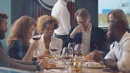 Waiter in protective face shield and mask serving food for diverse group of people in restaurant