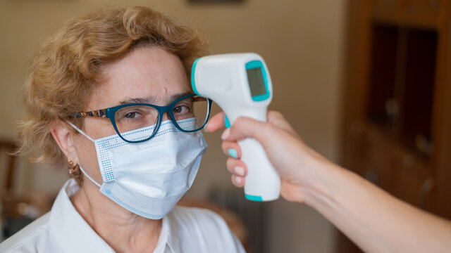 A Doctor Measures The Temperature Of An Elderly Woman Wearing A Mask With An Infrared Thermometer. House Call Medical Check-up