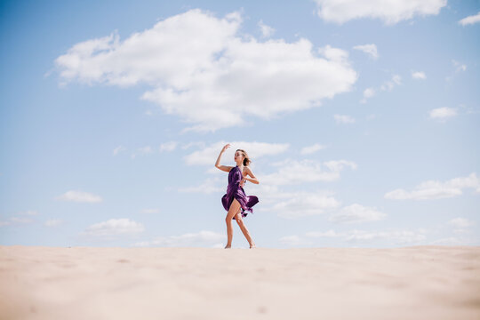 A young, slender girl in a beige dress with purple cloth in her hands posing in the desert in the wind