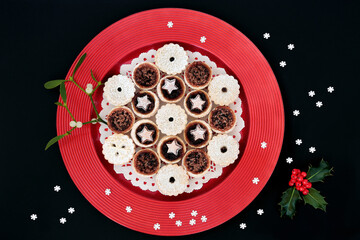 Homemade mince pies for Christmas on a red plate with winter berry holly, mistletoe & decorative snowflakes on black background. Festive food composition for the xmas holiday season. Flat lay, top vi