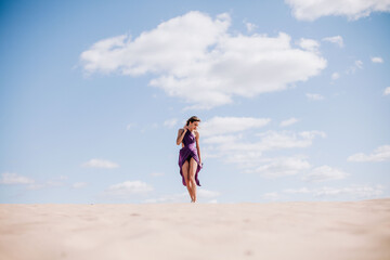 Fototapeta na wymiar A young, slender girl in a beige dress with purple cloth in her hands posing in the desert in the wind