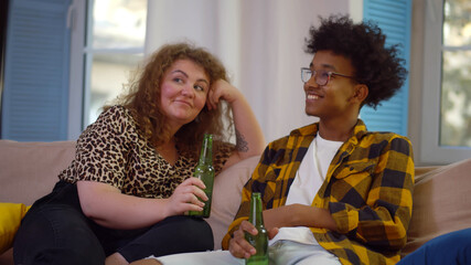 Mixed-race couple watching tv at home and drinking beer while sitting on sofa in cozy apartment