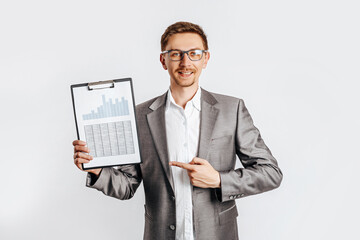 Young handsome brunet man in glasses in suit smiles and points at business with documents and graphs on white isolated background
