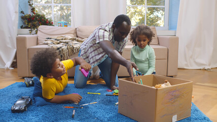 African father and two preschool children sitting on carpet and putting toys in box