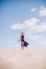 Fototapeta premium A young, slender girl in a beige dress with purple cloth in her hands posing in the desert in the wind
