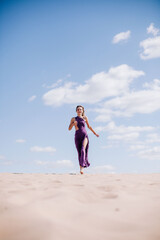 A young, slender girl in a beige dress with purple cloth in her hands posing in the desert in the wind
