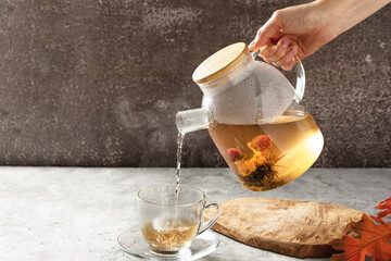 Woman hand pouring hot blossoming tea from glass teapot into a cup.