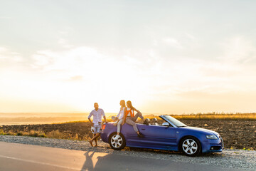 Group of friends having fun in convertible car during road trip at sunset. Young travel people driving a cabriolet during summer holidays. Happiness, vacation and youth lifestyle concept