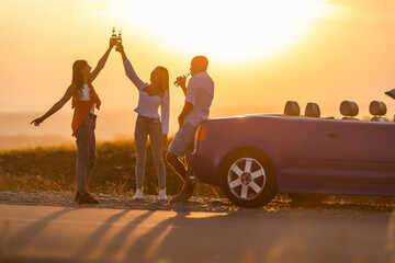 Group of happy young people with drinks in a car at sunset in summer.