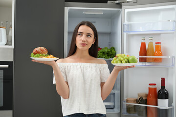 Woman choosing between fruits and burger with French fries near refrigerator in kitchen