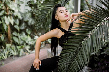 Fashion tropical portrait. Young attractive Asian woman model in trendy black overalls, with ponytail hair, sitting near green palm leaves, while posing in beautiful greenhouse wth tropical plants