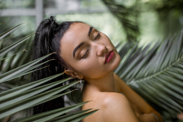 Asian woman model in tropical greenhouse. The portrait of mixed raced female model with dark long hair, posing among the leaves of beautiful tropical palm tree with eyes closed. Beauty concept