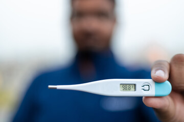 Sick man holds digital thermometer with body temperature reading in Fahrenheit up close to the camera.
