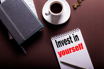 On a brown background, diaries, a white cup of coffee and a notebook with INVEST IN YOURSELF