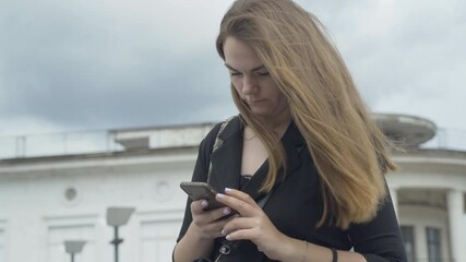 Concentrated Caucasian woman typing message on smartphone outdoors. Portrait of young beautiful brunette lady standing on urban street on windy day and messaging online.