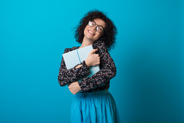Fototapeta premium Cute curly haired woman is embracing a present and smile on blue studio wall wearing glasses and dress