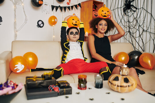 Funny brother and sister with Halloween make-up sitting on sofa in decorated room and posing with Jack-o-lanterns on their heads