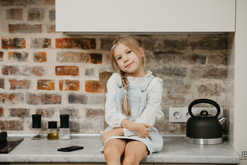 A blonde young girl with pale skin is sitting on the table in the kitchen.
