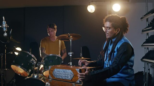 Music studio. Band rehearsal. Caucasian man playing drums, indian man playing harmonium. Talented musicians record their ideas.