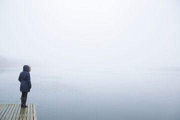 Young adult woman standing alone on edge of footbridge and staring at lake. Mist over water. Foggy...