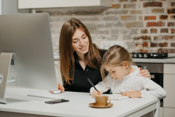 A young mom is hugging a daughter while she is drawing in the notebook at home. A gorgeous mother is looking at her blonde child which is doing homework.