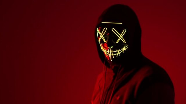 Anonymous shakes his head, halloween mask, unknown horror movie character, masked villain. A man in a mask in neon light on a red background. A man in a mask in neon light on a red background