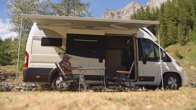 Caucasian Men Browsing Internet Outside of His RV Motorhome on a Camping. Scenic Camping Pitch.