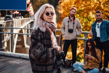 Well dressed beautiful blond posing with camera staying in foreground in background of autumn nature and her friends in daytime.