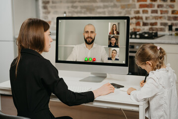 A young mother during a video conference with colleagues on the desktop computer while her daughter is writing in a notebook. A gorgeous mom is working remotely with her blonde child at home