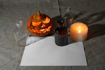 Carved pumpkin lantern with candle, chemical bottles and sheet of paper on gray linen cloth.