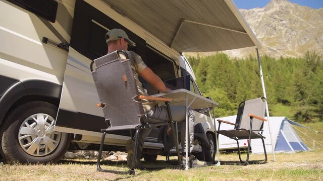 Men Using Internet on His Laptop Computer Next to his Motorhome Class B Camper Van, While on Camping in Some Remote Place Between Mountains