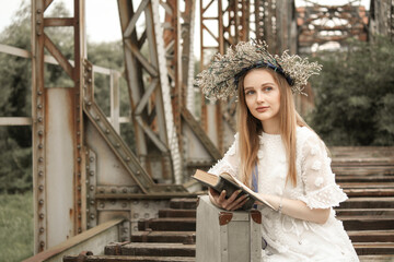 Woman dressed in a white dress holding a book and wearing a lei using her briefcase as a stand on a railway bridge with the bridge in a background